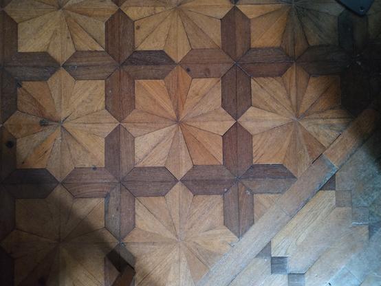 Top-down photo of wooden flooring. Most of the picture has dark brown crosses and light brown eight-pointed stars. The bottom right has light brown elongated hexagons and dark brown squares. A blueish light comes from the bottom right. 
The eight-pointed stars are not like the common octagrams of two overlapping squares of equal size. Instead, the vertices are on a square grid, presumably to simplify the production of the wooden tiles.