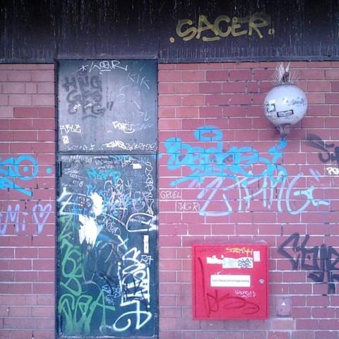 Mit Graffiti übersähte Tür in der Ruine des Ihmezentrums in Hannover-Linden. Rechts von der Tür ein feuerwehrroter Kasten für die Löschwassereinspeisung und eine an der Wand befestigte kugelförmige Laterne im Geschmack der Siebziger Jahre. Über der Tür spitze Nägel zur Taubenabwehr, darüber hat ein ehrenamtliches Wandlackierery das lateinische Adjektiv »sacer« gesprüht, das gleichermaßen heilig wie auch verrucht bedeuten kann.
