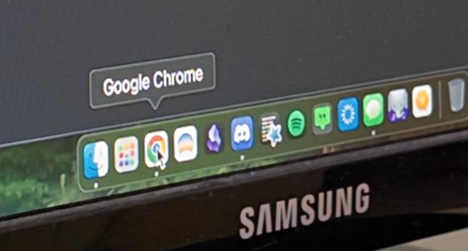 Photograph of a computer screen showing the macOS dock with a cursor hovering over the Google Chrome icon, which has a speech bubble above it that reads, "Google Chrome." The Samsung logo is visible on the bottom of the screen.