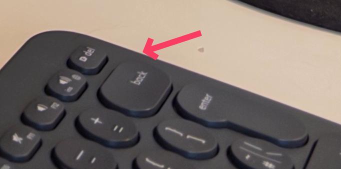 Photograph of a portion of a dark gray keyboard with a red arrow pointing to the "back" key. Other keys visible include "del", volume controls, "+ =", and "enter".