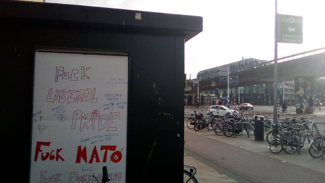 zie caption en alt bij andere foto. geparkeerde fietsen, paal met reclame hoog, viaduct en gebouw op achtergrond, langs weg met auto's. Op de viaduct paal staat een mozaiek achtige kleuring