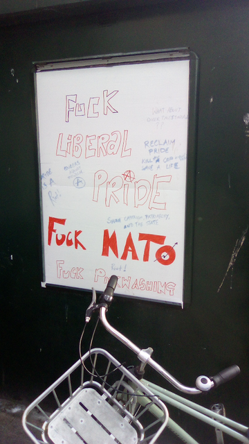 zwarte muur met wit gemaakt reclame bord. in open rood omlijnde letters: fuck liberal pride met anarchist A. Fcku nato in dicht gemaakte rode letters. In blauw: pride is a riot, reclaim pride, 
Queers against zionism, ... capitalism, patriarchy and the state

In open rood omllijnde letters fuck pinkwashing