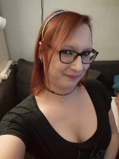 Das Bild zeigt eine Frau mit roten Haaren, die ein schwarzes T-Shirt trägt. Sie hat einn Haarreif mit blauen und grünen Akzenten auf dem Kopf und trägt eine schwarze Choker-Kette mit einem silbernen Anhänger. Ihre Haare sind mittellang und fallen ihr über die Schultern. Sie trägt eine Brille mit schwarzen Rahmen und hat ein sanftes Lächeln auf den Lippen. Im Hintergrund ist ein dunkles Sofa mit einem Teppich und ein Teil einer Wand mit einem weißen Stecker zu sehen. Die Frau scheint in einem Wohnraum zu sein.