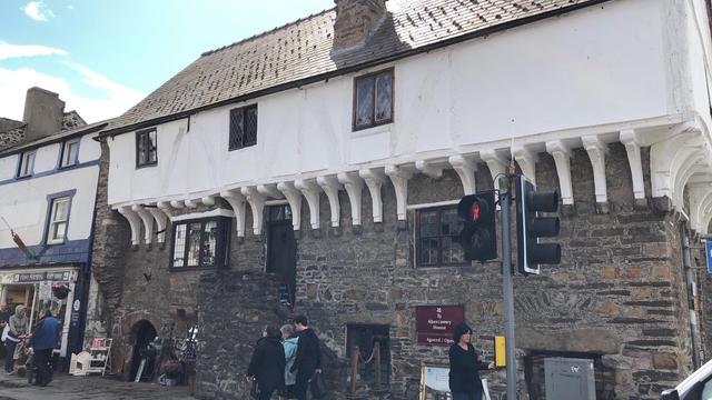 This image depicts Aberconwy House, a historic medieval merchant's house located in Conwy, Wales. The building is constructed with stone on the lower level and features white timber framing with exposed beams on the upper level. The roof is steeply pitched and covered with slate tiles. The upper story slightly overhangs the lower part, supported by prominent wooden brackets.

In front of the building, there is a pedestrian crossing with a traffic light showing a red signal for pedestrians. Several people are walking on the cobblestone pavement, and there are shops adjacent to Aberconwy House. The weather appears clear with blue skies and scattered clouds, suggesting a pleasant day.