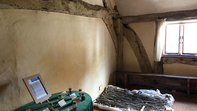 This picture depicts an interior of Aberconwy House, characterized by its rustic, exposed wooden beams and plastered walls. There's a wooden-framed window on the right side, partially covered by a light curtain, allowing natural light to stream into the room.

In the foreground, there's a table covered with a green cloth, displaying small jars labeled with handwritten tags, possibly containing herbs, spices, or other historical artifacts. A framed informational sign is also present on the table, providing context about the display.

On the floor near the window, there is a bundle of sticks arranged neatly on a white cloth or padding, suggesting it might be part of an exhibit or demonstration related to historical living conditions or practices. A simple wooden bench runs beneath the window, adding to the room's minimalistic and historical aesthetic.