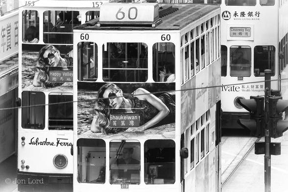 This is a black and white street photo in landscape format. Dominating the image Is an elevated view of three vintage Hong Kong trams. The closest tram is in the centre of the frame and moving towards the camera. The driver can be seen sitting in the cab in the centre of the tram. Above is the sign indicating the destination: Shaukiwan and 筲箕灣 in Chinese characters. There is an advert on the front with a large photo of a young woman lying on the ground with long hair and sun glasses, though the product remains unknown. Passengers can be seen on the upper deck and many of the windows are open. In the upper right corner is a further tram again moving towards the camera, this one with its destination: Causeway Bay, 銅鑼灣. Along the left margin is the last tram, this one moving away from the camera, its destination: Happy Valley, 跑馬地.
The Location: Causeway Bay, Hong Kong (2012).