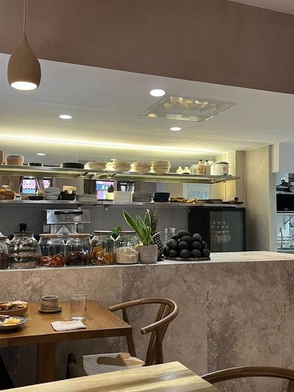 The image shows a restaurant or cafe interior with a counter displaying jars, plants, and a pile of avocados. A wooden table and chairs are in the foreground.