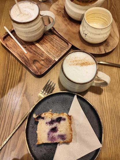 A table setting with a blueberry cake on a black plate, a fork, and two mugs of foamy drinks. There's also a teapot and a cup on a wooden tray.