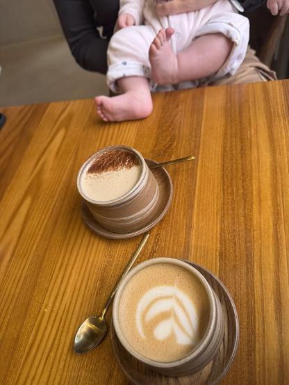 Two cups of coffee on a wooden table, with a baby's legs and feet visible in the background.
