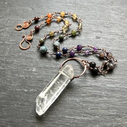 A photo of a large, wire-wrapped copper link statement necklace with a rainbow array of semi-precious stone beads and a beautiful raw quartz crystal pendant on a dark grey slate background.  The quartz crystal point focal itself is translucent and topped with a rustic, hammered copper halo setting. The hook and eye clasp is also hand forged in copper.
