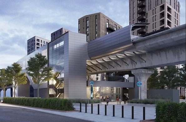 An architectural rendering of a contemporary transport station with a sleek, grey exterior. The station features large glass windows and is situated beneath an elevated track. Surrounding the station are modern residential buildings, and a pathway lined with trees leads to the entrance where people are walking.

TfL Graphic - Artist's Impression of Pontoon Dock 2