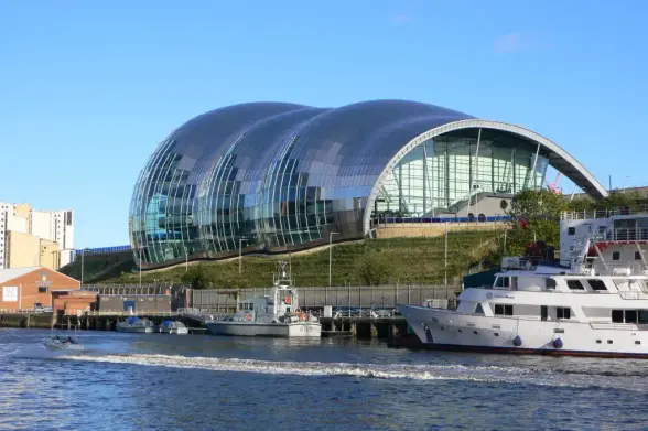 The image features a distinctive modern building with a unique architectural design. It has a curved, glass exterior that resembles large waves or shells. The building is situated near a body of water, with boats visible in the foreground. The surrounding area appears to include some greenery and other structures. This style of architecture is often associated with cultural or performance venues.

Image Credits: Wikimedia / Jimfbleak at English Wikipedia / CC BY-SA 3.0