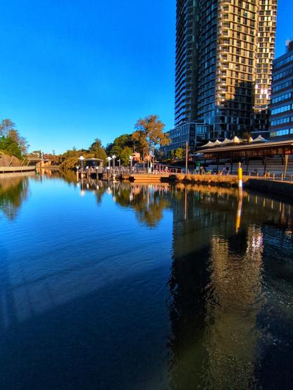 The turning basin for the Parramatta RiverCat ferry service. On the left is a concrete seating area underneath a wooden structure. A wharf, where passengers embark and disembark, floats on the water. A tall apartment skyscraper towers overhead. The sky is very blue, reflected in the water. There's trees on the riverbank in the distance.