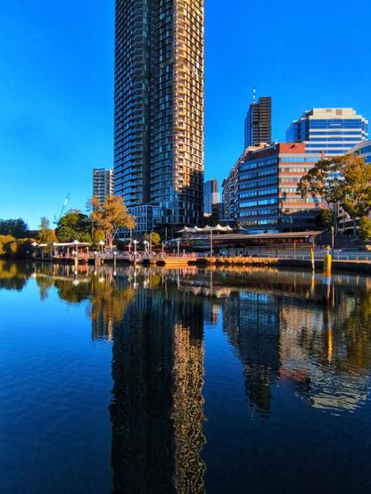 The same scene as before, shot further around the turning basin. The reflection of the tall apartment skyscraper is now clearly visible in the blue river water. Some more office buildings are now visible to the left of the wooden seating structure.