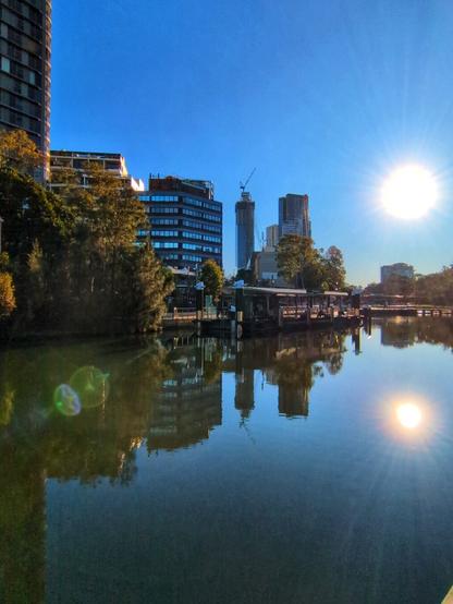 Looking at the Quay from the opposite side, slightly downriver. The sun looks white in this photo against a very blue sky. The water is teal, reflecting a mix of trees and sky. More office buildings are visible on the horizon.