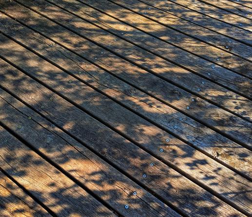 A high-angle shot looks down onto a sun-drenched wooden deck, composed of parallel planks fastened with visible screws. Diagonal shadows, cast by what appear to be leaves or branches, create a dappled pattern across the warm, natural brown tones of the wood. The texture of the planks, with their grain and slight imperfections, is clearly visible, suggesting an outdoor setting under natural light.