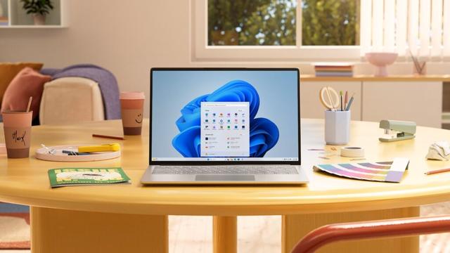 A laptop displaying the Windows 11 home screen is placed on a round wooden table in a cozy, well-lit room. The table is surrounded by various items including two coffee cups, one labeled "Mark," a tray with stationery, a magazine titled "ISSUE N. 04 Women in Tech," a color palette, a stapler, and a pencil. The background features a window with a view of greenery, a couch with cushions, and a shelf with a plant. The image highlights a modern, comfortable workspace setup with the latest Windows operating system.