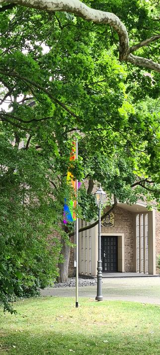 Etwas verdeckt durch einen Laubbaum flattert eine Regenbogenfahne im Wind. Dahinter das Portal einer Backsteinkirche mit einer kurzen Vorhalle mit jeweils drei schmalen Fenstern auf jeder Seite. Über der Tür ein Mosaik, das den hl. Albertus Magnus darstellt.