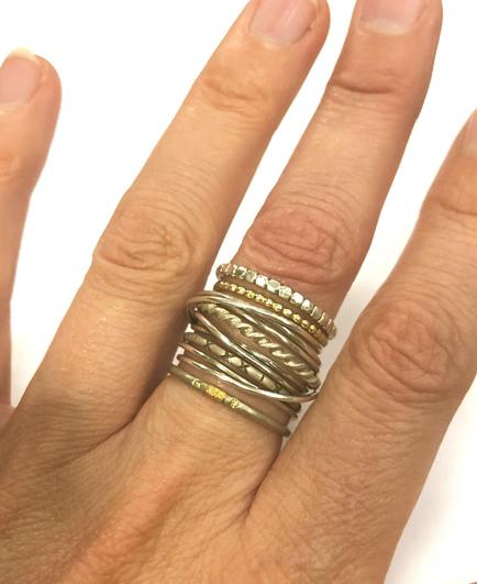 A hand wearing multiple rings, featuring a mix of silver and gold tones, with various textures and designs. The background is plain and white.