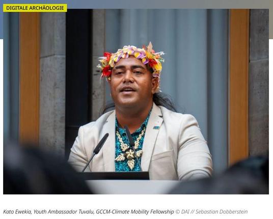 Ein Mann mit traditioneller Blumenkopfbedeckung spricht am Rednerpult. Er trägt ein beige-farbenes Jackett über einem gemusterten türkisfarbenen Hemd und eine Muschelkette. Bildunterschrift: Kato Ewekia, Youth Ambassador Tuvalu, GCCM-Climate Mobility Fellowship.