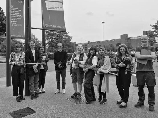 Image: black-and-white photos of presumably all the artists in the exhibition, smiling towards the camera outside RUA RED • “Exhibiting Artists: David Beattie, Ala Buisir, Cecilia Bullo, Pauline Cummins, Lauren Kelly, Maria McKinney, and Fiona Whelan    Together in Commune, is the first exhibition of Rua Red’s Studio Programme, curated by Marysia Więckiewicz and featuring work by Rua Red’s current resident studio artists: David Beattie, Ala Buisir, Cecilia Bullo, Pauline Cummins, Lauren Kelly, Maria McKinney, and Fiona Whelan.    This exhibition marks an important moment for Rua Red, highlighting the depth and breadth of the practices nurtured and supported within these walls. Working closely with the curator in the months leading up to the exhibition, each artist presents work that reflects their individual practice, while collectively exploring themes central to socially engaged contemporary art.…”