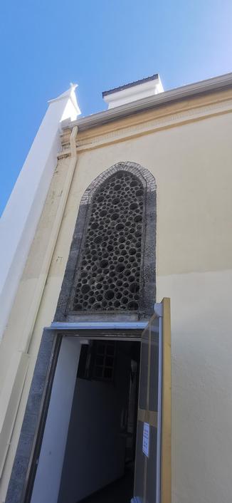 Photograph of the side entrance of a church in Réunion Island. Above the entrance, dozens of circular openings of various diameter (from 5 to 15cm) arranged in the shape of a tall arch. From a distance, it looks like a honeycomb, or the interior of a sponge.