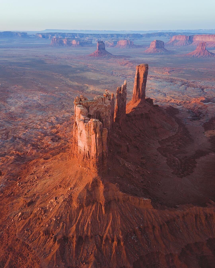 On observe ici un vaste désert de terre rouge, sculpté par l’érosion depuis des millions d’années. Au premier plan, une formation rocheuse élancée se dresse majestueusement, illuminée par la lumière dorée du lever ou coucher du soleil. À l’horizon, d’autres mesas et buttes s’étendent à perte de vue, donnant une impression d’infini. Le paysage est emblématique du Far West américain et a souvent servi de décor pour des films westerns classiques.