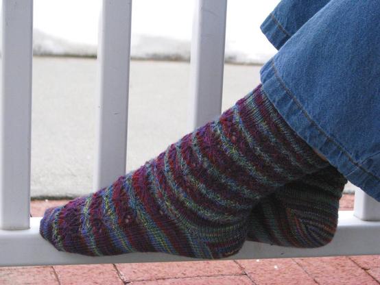 a pair of feet stretched out and resting against a railing with snow in the background, the feet are wearing a pair of purple socks with a cables and a stitch pattern resembling butterflies or abstract flowers.