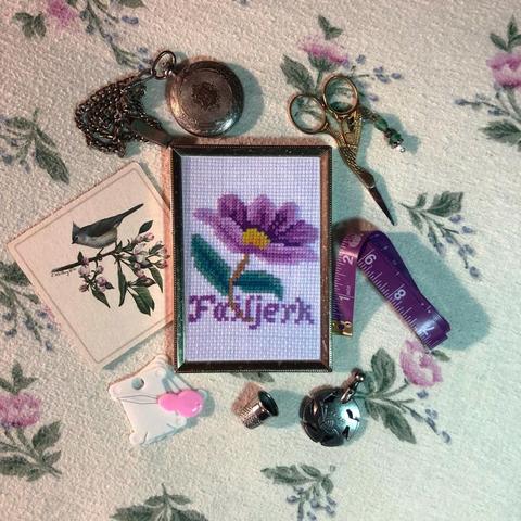 a small piece of cross stitch inside a thin gold frame surrounded by various bits of vintage looking sewing accouterments. the cross stitch is of a purple daisy like flower with the word "Failjerk" written below it in matching purple script.