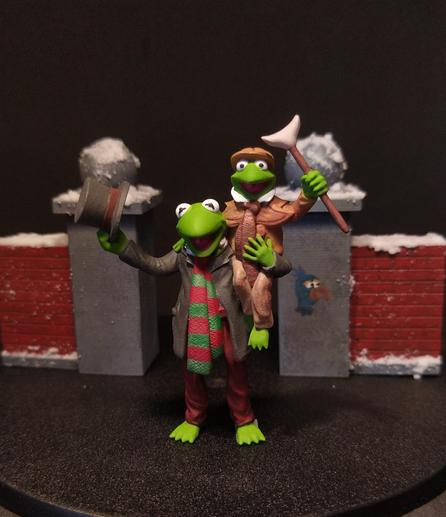Das Bild zeigt zwei Figuren, die nach dem Stil der Muppets gestaltet sind, vor einem winterlichen Hintergrund. Die Figur im Vordergrund trägt einen grauen Anzug, eine rote Hose und einen grünen und roten Streifenmuster-Schal. Sie hält einen schwarzen Zylinder in der Hand und lächelt. Die andere Figur, die auf der ersten Figur sitzt, trägt einen braunen Anzug, eine braune Mütze und hält einen Schaufelkopf in der Hand. Beide Figuren sind grün und haben große Augen. Der Hintergrund zeigt eine winterliche Szene mit Schnee auf den Dächern und einer roten Ziegelmauer. Ein kleines Bild eines blauen Vogels ist an der Wand zu sehen.