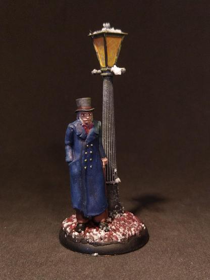 Das Bild zeigt eine detaillierte Miniaturfigur eines älteren Mannes, der in einem blauen Mantel und einem hohen Zylinderhut steht. Er hält einen Stock in der rechten Hand und steht neben einer klassischen Straßenlaterne, die mit Schnee bedeckt ist. Die Laterne hat ein gelbes Licht und ist auf einem schwarzen Pfosten montiert. Der Boden ist mit Schnee und rotem Erdboden bedeckt, was eine winterliche Szene schafft. Die Miniaturfigur und die Laterne sind auf einem runden Sockel platziert, der die Szene abgrenzt. Die Farben sind sorgfältig gewählt, um eine realistische Darstellung zu erzielen.
