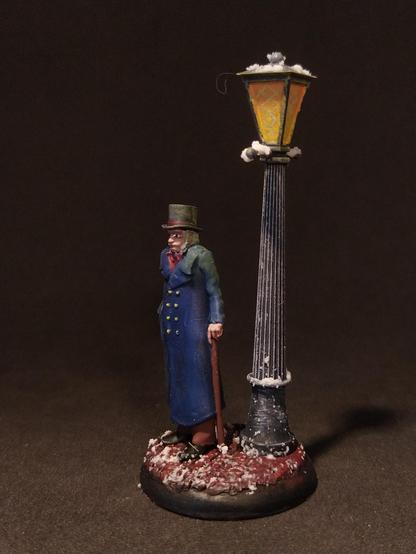 Das Bild zeigt eine detaillierte Miniaturfigur eines älteren Mannes, der in einem blauen Mantel und einem hohen Zylinderhut steht. Er hält einen Stock in der rechten Hand und steht neben einer klassischen Straßenlaterne, die mit Schnee bedeckt ist. Die Laterne hat ein gelbes Licht und ist auf einem schwarzen Pfosten montiert. Der Boden ist mit Schnee und rotem Erdboden bedeckt, was eine winterliche Szene schafft. Die Miniaturfigur und die Laterne sind auf einem runden Sockel platziert, der die Szene abgrenzt. Die Farben sind sorgfältig gewählt, um eine realistische Darstellung zu erzielen.