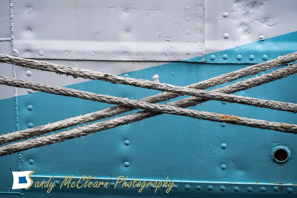 Two sets of two mooring ropes crossing against the background of a riveted steel plate ship hull.