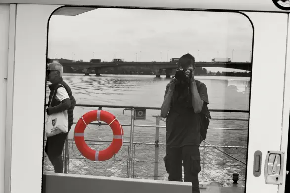 Reflet d'un fleuve et d'un pont dans la fenêtre d'un bateau. Le photographe et une autre personne, au bord du cadre de la fenêtre se reflètent également. Photo noir et blanc, avec la bouée de sauvetage en couleur.