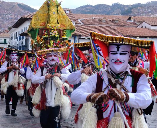 Photograph of an extraordinary procession in Peru. The figures are heading diagonally past the camera, and are dressed identically: masks reminiscent of that of the protagonist in V for Vendetta (which was a white mask with a pointed chin and small goatee with a striking moustache, with a smiling but intimidating expression), with white shirts and black waistcoats. the hats are unusual, being flat, wide and horizontal with tasselled fronts and ribbons hanging from the side. They are carrying white pompoms strung from their clothing. 

In the middle of the procession is a figure of the Virgin Mary carried aloft, dressed in a fabulous green and gold robe. Unfortunately, her head is not visible as the photographer cut it out.