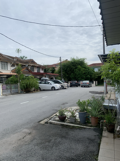 The image depicts a residential street scene. In the foreground, there is a paved road with a few parked cars, including a white sedan and a silver hatchback. The road is flanked by houses with red-tiled roofs, some with visible satellite dishes. The houses have a mix of architectural styles, with one featuring a covered carport. On the right side, there is a sidewalk with potted plants, including a variety of greenery in different pots. The sky is overcast, suggesting a cloudy day. Power lines run across the top of the image, and the overall atmosphere is calm and quiet, typical of a suburban neighborhood.

Provided by @altbot, generated privately and locally using Ovis2-8B

🌱 Energy used: 0.188 Wh