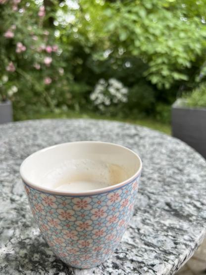 Ein geblümter Kaffeebecher auf einem Bistrotisch.
Im Hintergrund eine rosafarbene Kletterrose und eine weiße Girlandenhortensie