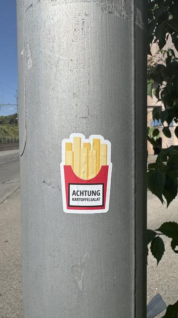 Sticker on an electric pole representing a portion of French fries in a red box. The tag on the box reads « achtung kartoffelsalat »
