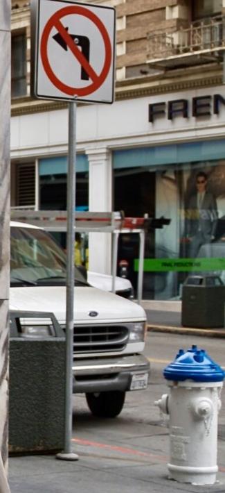 Photo showing the front of a workers white van and behind it is another partial van with a ladder on its roof. Two concrete waste cans and a fire hydrant are in the frame. In the foreground is a No Left Turn sign.