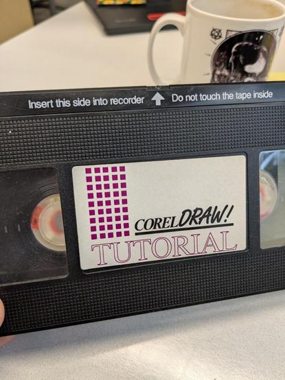 A photograph of a VHS casette tape that says "Corel Draw! Tutorial" on the label. The top of the VHS says "Insert this side into recorder, do not touch the tape inside".

In the background you can see a blurry coffee mug and my desk.