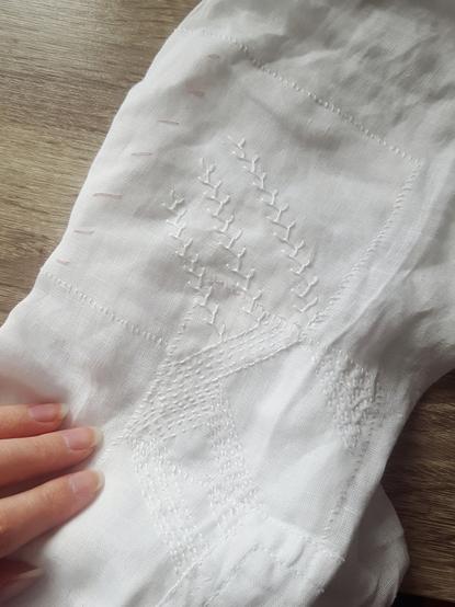Overlooking a white linen shirt sleeve that has a number of overlapping patches on it. From the wrong side they've been whipstitched on, and on the right side this shows up as a line of slanted spaced stitches. On top of the patches there's topstitching or embroidery of varying style and care, going from diagonal running stitches to feather stitch. It's a little bit of a mess.