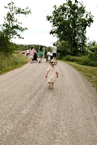A young child wearing a white dress and a flower crown walks alone on a gravel path towards a group of people, some pushing strollers, surrounded by green trees and grass on a cloudy day.