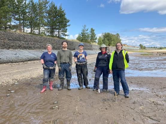 a group of 5 people, all a bit muddy standing on a shore at low tide beside some newly planted saltmarsh. They are all smiling. 
some of the protective fencing is visible in the distance to the right of the group