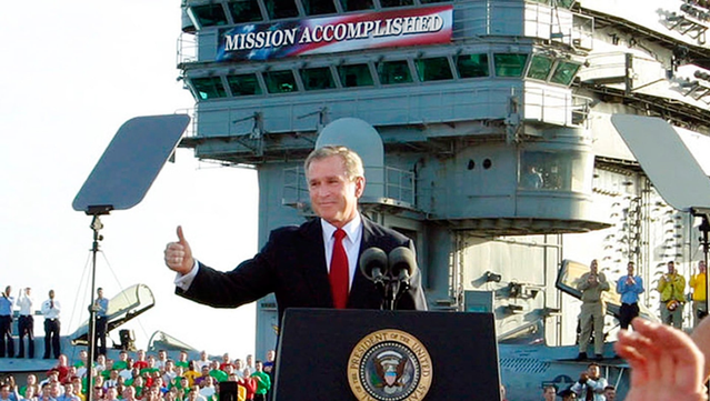George W. Bush standing in front of a Mission Accomplished banner on an aircraft carrier, giving a thumbs up from behind a presidential podium.