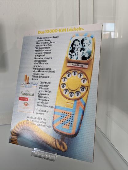 Im Grunde Werbung für Zigaretten.
Neben einer solchen Schachtel ein Bild von einen verkabelten Handteil eines Telefons in orange-blau mit Wählscheibe und kleinem Bildschirm.
