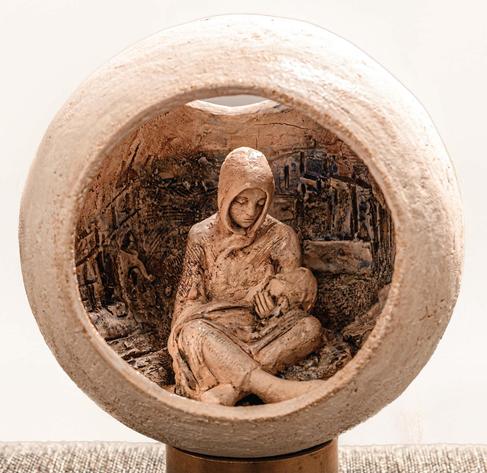 Image: Ann Meldon Hugh: Fragile World, ceramic and bronze, 13 x 13 x 24cm | photo of the artwork; we see a mother breastfeeding a child inside a sphere that is open at the front and above towards the back; the mother is wearing a hooded garment and is looking down at the child; the outside of the globe is rough in texture and off-white; the interior in which the mother is sitting is rougher still, and resembles a blackened cityscape • “An exhibition of new work by award winning sculptor Ann Meldon Hugh. A personal response to her environment and experience, the exhibition features 30 stunning works in ceramic and bronze capturing the beauty and fragility of our world.    Ann Meldon Hugh was born in Dublin in 1952 and graduated from the National College of Art and Design in Dublin in 1975, specialising in sculpture. She currently lives in Co. Meath. Ann has been working as a full-time sculptor since 1989 and has been commissioned to complete many public sculptures throughout Ireland…”