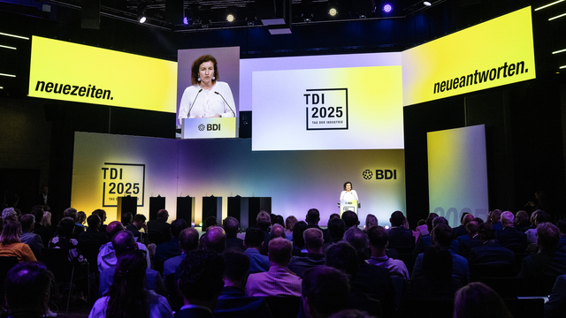 Konferenzraum mit Publikum und einer Präsentation der Veranstaltung TDI 2025. Große gelbe Banner mit den Worten 'neuezeiten.' und 'neueantworten.' sind zu sehen. Bundesforschungsministerin Dorothee Bär hält eine Rede.