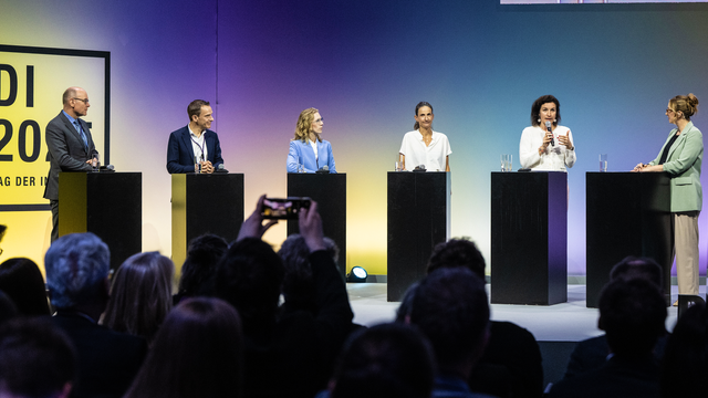 Diskussionsrunde mit sechs Personen auf einer Bühne vor einem Publikum. Drei Personen tragen Businesskleidung und stehen hinter Pulten mit Mikrofonen und Wassergläsern. Die Bühne hat einen blauen und gelben Hintergrund mit der Aufschrift 'ADI 2023 Tag der Industrie'.
