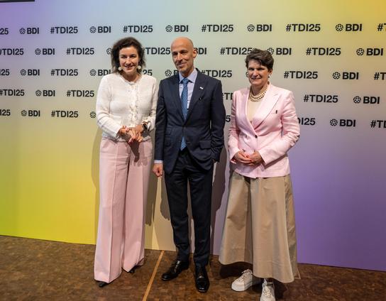Zwei Frauen und ein Mann stehen vor einem Hintergrund mit dem Hashtag #TDI25 und dem Logo des BDI. Die Frauen tragen elegante Kleidung in verschiedenen Rosa- und Beigetönen, während der Mann einen dunkelblauen Anzug mit Krawatte trägt.