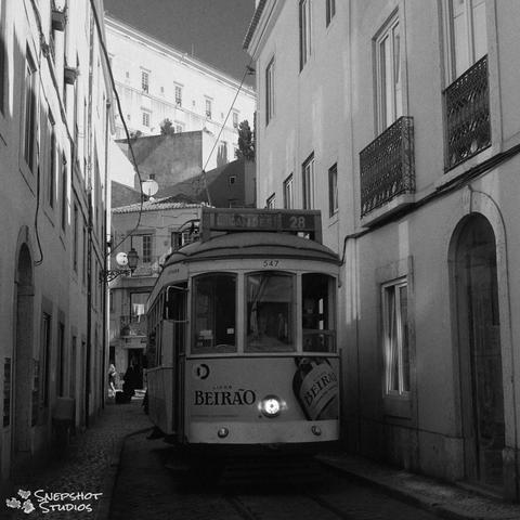 Square black and white photograph of an old streetcar with its light on coming through a narrow street